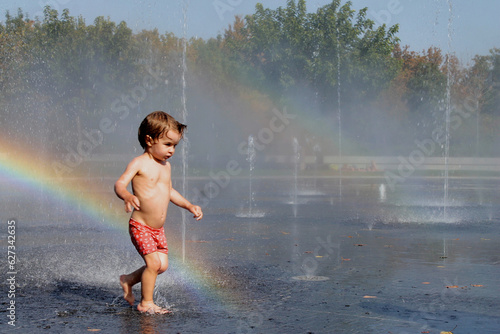 Toddler in a swimsuit playing in some water fountains in the city of Madrid, Spain with a rainbow behind