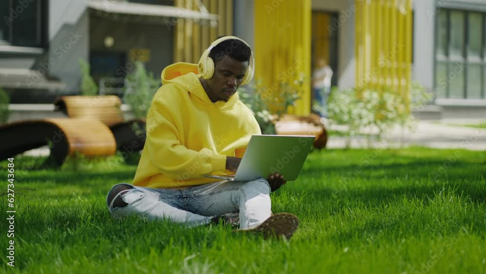 Young Black Guy Sitting On Grass In Park In Summer Day And Using Laptop With Wireless Headphones Young Black Guy Sitting On Grass In Park In Summer Day And Using Laptop With Wireless Headphones