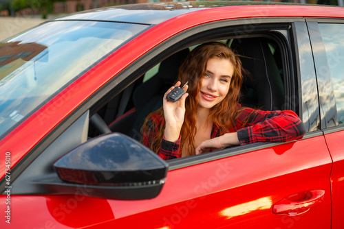 Smiling red-haired girl in a plaid shirt holding car keys while sitting in a red car