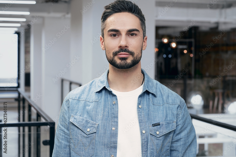 Naklejka premium Portrait of young bearded man standing in the office while posing at camera. Modern coworking on the background. Startup, coworking, management concept