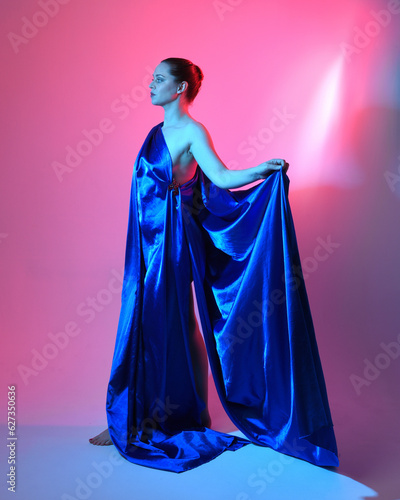 Full length portrait of beautiful female model wearing elegant flowing toga gown of blue silk drapery, isolated on studio background with cinematic colourful lighting.