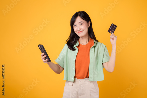 Fotografia Embrace the ease of mobile shopping with an enchanting young Asian woman in her 30s, wearing orange shirt and green jumper, using smartphone while displaying credit card on yellow studio background