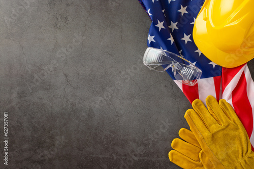 Celebrating the tireless efforts of construction workers on Labor Day. Top-down image showcasing flag, helmet, gloves and goggles on grunge textured grey concrete. Copyspace available for ads or text