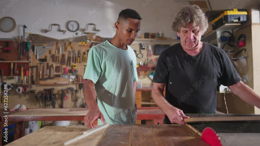 Carpenter and apprentice working at workshop, teacher orienting young man with saw machine at carpentry. Wood sawing scene of mentor teaching student Carpenter and apprentice working at workshop, teacher orienting young man with saw machine at carpentry. Wood sawing scene of mentor teaching student