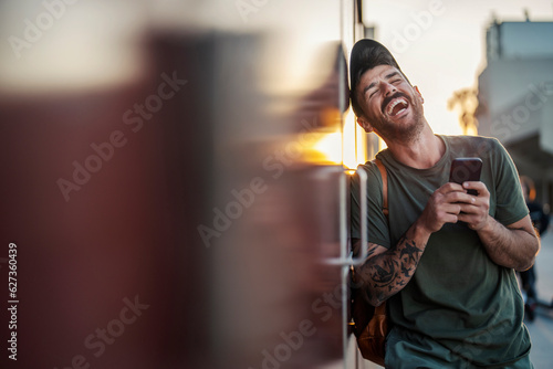 A tattooed man is leaning on the wall on the street and laughing while holding a phone.