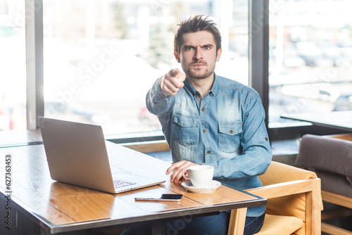 Fotografie Portrait of strict bossy young bearded handsome man freelancer in blue jeans shirt working on laptop, pointing at camera, selecting you