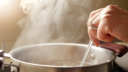 Female hand stirring boiling soup in metal pan with spoon. Vapour slowly rising from hot water.