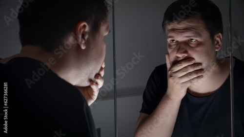 Upset and stressed man standing at mirror and covering face with hands. Concept of depression, suicide, stress, mental illness, loneliness and frustration.