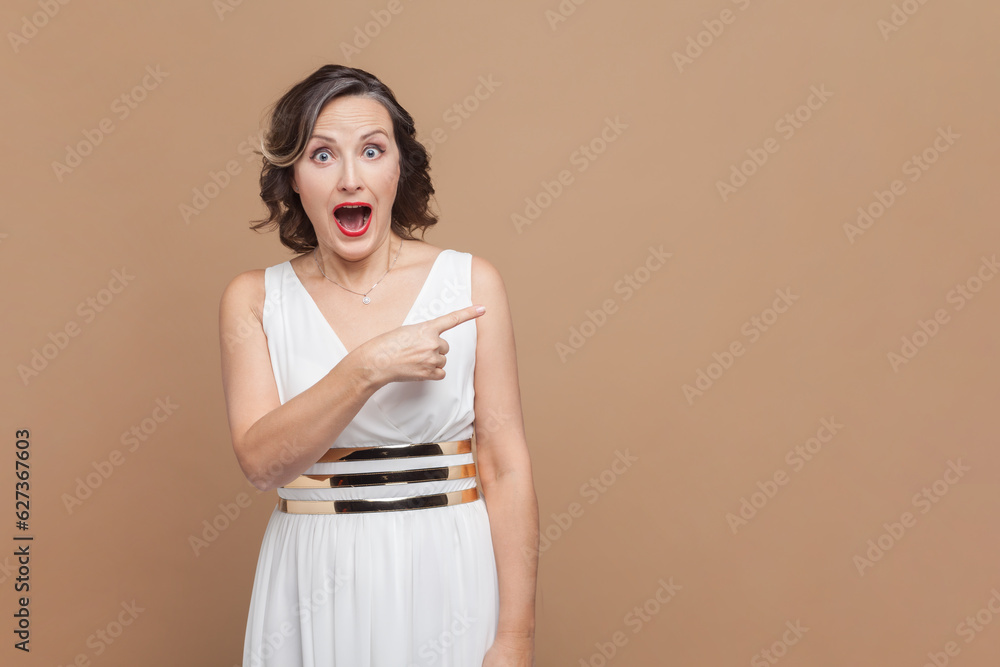 Portrait of happy joyful middle aged woman with wavy hair pointing away at copy space for advertisement, wearing white dress. Indoor studio shot isolated on light brown background. Portrait of happy joyful middle aged woman with wavy hair pointing away at copy space for advertisement, wearing white dress. Indoor studio shot isolated on light brown background.