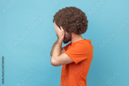 Side view of man with Afro hairstyle in orange T-shirt closing eyes with hand, dont want to see that ignoring problems, hiding from stressful situations. Indoor studio shot isolated on blue background