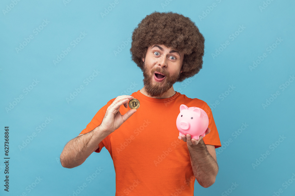 Portrait of man with Afro hairstyle wearing orange T-shirt holding piggy bank and golden bitcoin, looking at camera with shocked expression. Indoor studio shot isolated on blue background. Portrait of man with Afro hairstyle wearing orange T-shirt holding piggy bank and golden bitcoin, looking at camera with shocked expression. Indoor studio shot isolated on blue background.
