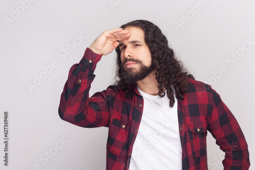 Portrait of man with long curly hair in checkered red shirt looking far away at distance with hand over head, sees something with attentive expression. Indoor studio shot isolated on gray background.