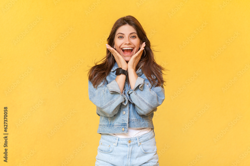 Portrait of excited extremely happy woman in denim jacket standing with palms near face, screaming happily, rejoicing her victory, screaming hurray. Indoor studio shot isolated on yellow background. Portrait of excited extremely happy woman in denim jacket standing with palms near face, screaming happily, rejoicing her victory, screaming hurray. Indoor studio shot isolated on yellow background.