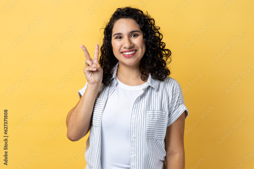 Portrait of happy optimistic woman with dark wavy hair doing victory gesture and looking smiling to camera, showing peace, v sign with double fingers. Indoor studio shot isolated on yellow background. Portrait of happy optimistic woman with dark wavy hair doing victory gesture and looking smiling to camera, showing peace, v sign with double fingers. Indoor studio shot isolated on yellow background.