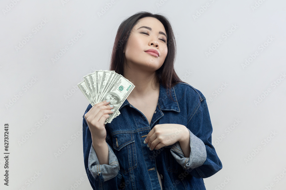 Portrait of satisfied rich proud brunette woman in blue denim jacket standing holding big fan of dollars, gets salary or winning jackpot. Indoor studio shot isolated on gray background. Portrait of satisfied rich proud brunette woman in blue denim jacket standing holding big fan of dollars, gets salary or winning jackpot. Indoor studio shot isolated on gray background.