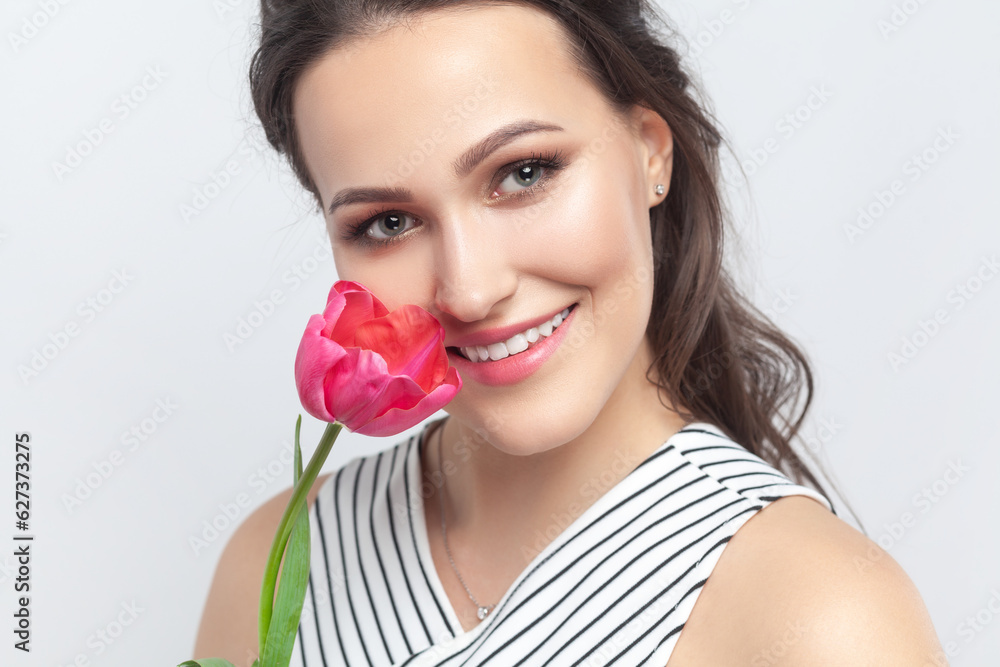 Closeup portrait of romantic smiling joyful brunette woman standing looking at camera, holding pink tulip, enjoying flower, wearing striped dress. Indoor studio shot isolated on gray background. Closeup portrait of romantic smiling joyful brunette woman standing looking at camera, holding pink tulip, enjoying flower, wearing striped dress. Indoor studio shot isolated on gray background.