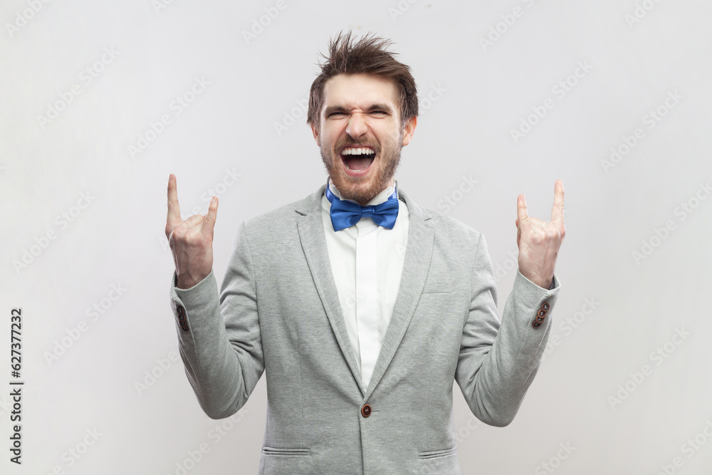 Portrait of crazy funny excited handsome bearded man standing with rock and roll gesture, screaming with excitement, wearing grey suit and blue bow tie. Indoor studio shot isolated on gray background. Portrait of crazy funny excited handsome bearded man standing with rock and roll gesture, screaming with excitement, wearing grey suit and blue bow tie. Indoor studio shot isolated on gray background.