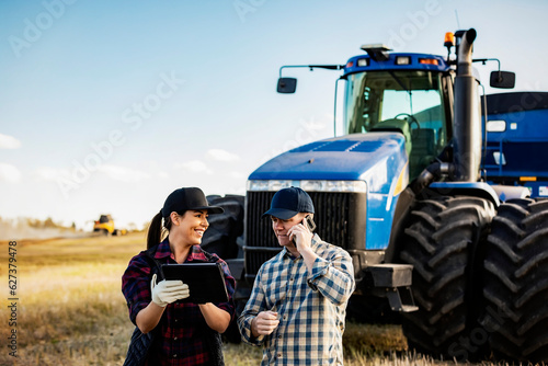 A husband and wife using portable wireless devices to manage and monitor their canola harvest and making a call while standing in front of a big tractor; Alcomdale, Alberta, Canada