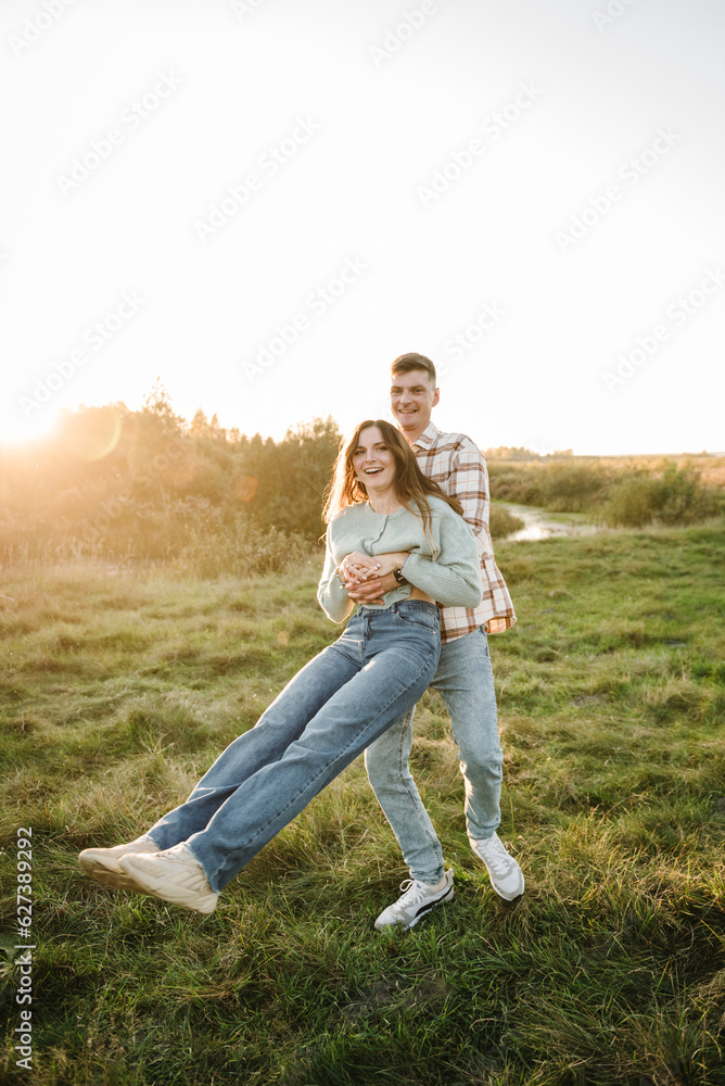 Young man spins woman, walking spending time together in nature. Couple hugging and smiling in grass in field at sunset. Family holiday outdoors. Female embrace male in summer park. Sunset scene. Young man spins woman, walking spending time together in nature. Couple hugging and smiling in grass in field at sunset. Family holiday outdoors. Female embrace male in summer park. Sunset scene.