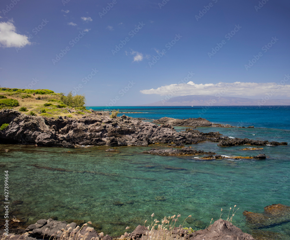 Close-up of the clear, turquoise water along the rocky shore with distant view of a boat moored on the horizon and silhouetted mountains under a blue sky; Maui, Hawaii, United States of America Close-up of the clear, turquoise water along the rocky shore with distant view of a boat moored on the horizon and silhouetted mountains under a blue sky; Maui, Hawaii, United States of America