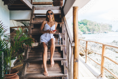 Happy woman sitting on stairs of villa with cellphone