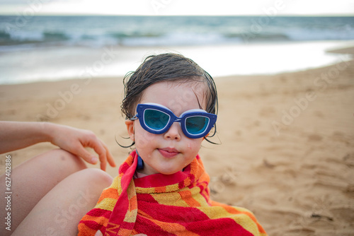 Close-up portrait of young girl wearing swimming goggles warpped in a beach towel and sitting by her mother on the beach; Maui, Hawaii, United States of America photo