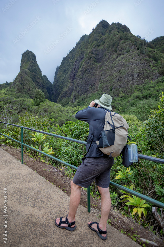 View taken from behind of a man standing on a walkway photographing the view of the Kuka‘emoku, Iao Needle in the lush, Iao Valley in Central Maui; Maui, Hawaii, United States of America