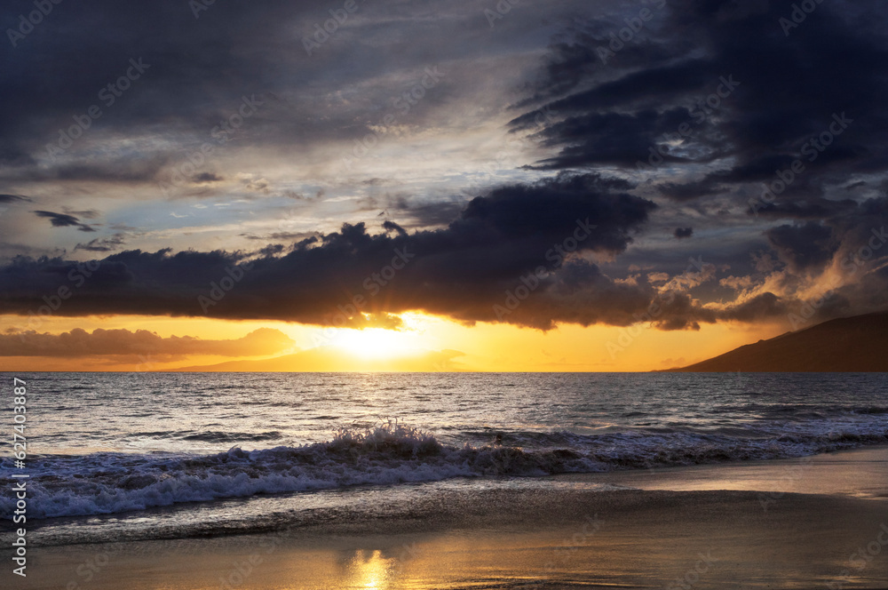 Ocean waves along the shoreline of Kamaole 2 Beach at twilight with a golden sun glowing under a cloudy sky; Kihei, Maui, Hawaii, United States of America