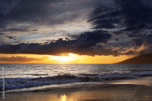 Ocean waves along the shoreline of Kamaole 2 Beach at twilight with a golden sun glowing under a cloudy sky; Kihei, Maui, Hawaii, United States of America