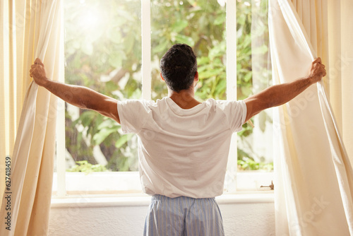 Back view, man and open curtain at window to good morning, sunrise and sunshine to relax in room at home. Guy, wake up and opening bedroom drapes in apartment for sunlight, fresh air or start new day photo