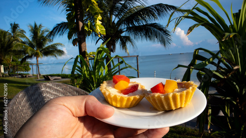 Male hand holding white plate with two slices of mini fruit pie, with slices of grapes, peaches and papaya on wicker table during breakfast in hotel, vacation with happy family 