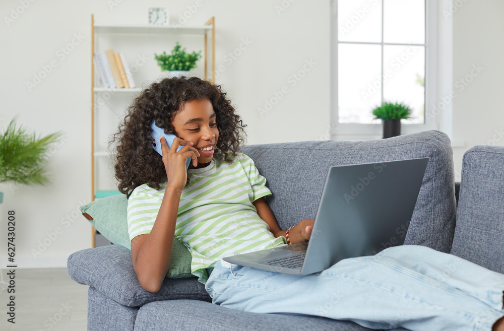 Cheerful African American teenage girl talking on mobile phone while lying with laptop on sofa. Curly-haired girl spends her free time in her room browsing social networks and talking with friends. Cheerful African American teenage girl talking on mobile phone while lying with laptop on sofa. Curly-haired girl spends her free time in her room browsing social networks and talking with friends.