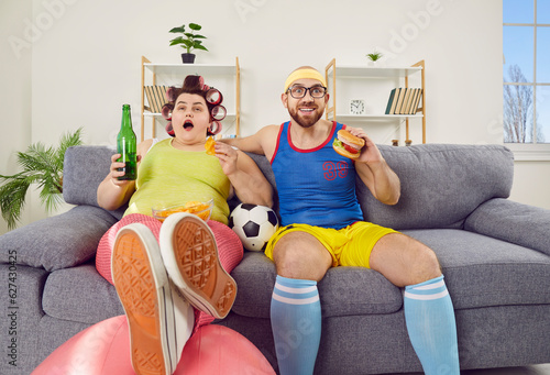 Funny family couple eating and watching football on TV on cheat day. Fat woman and thin man sitting on sofa with soccer ball, watching sports match and eating junk food. Unhealthy lifestyle concept