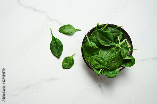 A bunch of Fresh baby spinach leaves in the in a black ceramic bowl on white marble background. Top view and flat lay photo with copy space