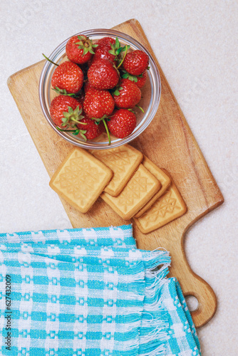 Fresh ripe strawberry in a glass bowl and cookies at wooden board on a light table. Heap of strawberries. Top view.
