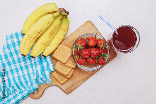 Glass bowl with strawberries, bananas, glass of red juice and cookies on light table. Top view.