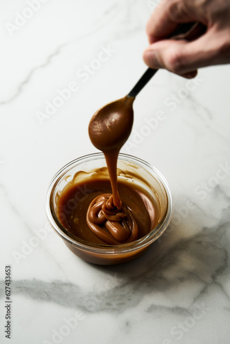 Salted Caramel sauce in glass bowl on white marble background. The hand holding the spoon dips into the caramel sauce, the caramel stretches. Butter, sugar with cream and salt. 