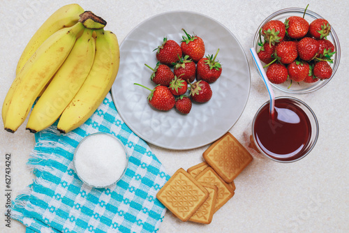 Plate and glass bowl with strawberries, banana branch, glass of red juice, bowl of sugar and cookies on light table with blue background. Top view.