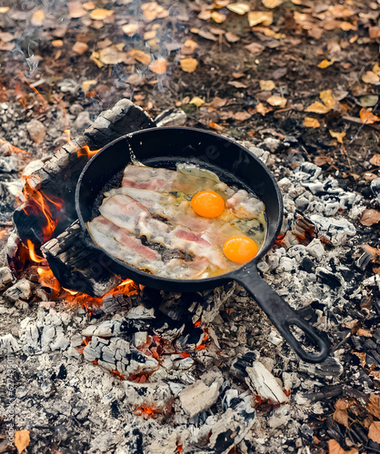 fried bacon and eggs in a pan over a campfire in the forest