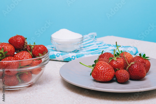 Heap of freshly harvested strawberries on a light table . Strawberries served in a ceramic plate and glass bowl with bowl of sugar.