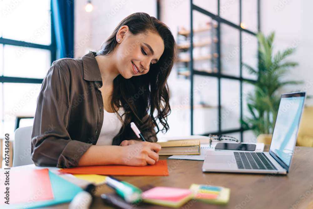 Smiling female student writing on colored paper while studying on laptop Smiling female student writing on colored paper while studying on laptop