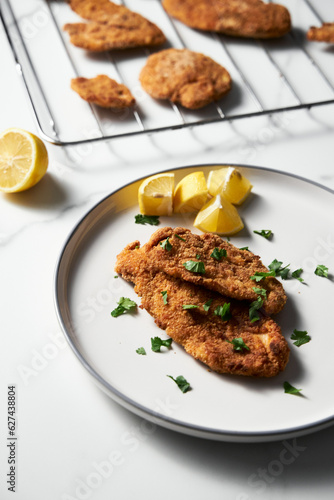 Baked Chicken Schnitzel on white plate with lemon. Rack or grid with baked schnitzel in breadcrumbs on background. 