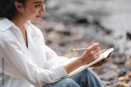 Photo of young attractive Asian woman writing on diary while sitting outdoors in the park  Writer sitting in garden  Female freelance are relaxing to writing story on jobs  beautiful Asian women