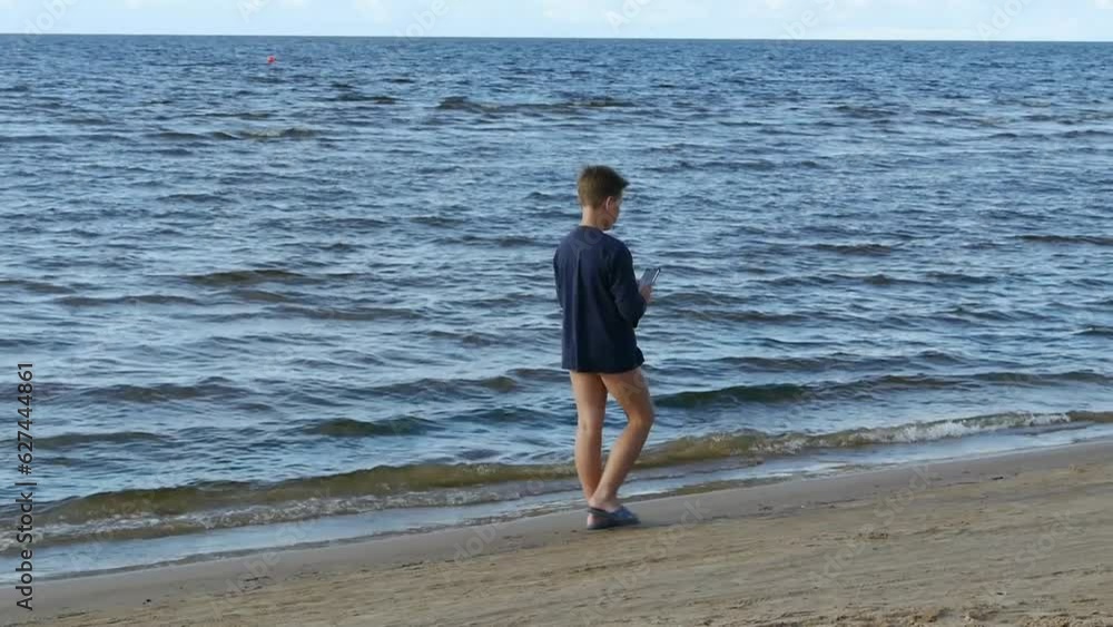 Caucasian boy walks along the beach and speaks from a smartphone. A 9-13 year old boy is walking along the Baltic Sea beach. The child man walks on the beach. Caucasian boy walks along the beach and speaks from a smartphone. A 9-13 year old boy is walking along the Baltic Sea beach. The child man walks on the beach.