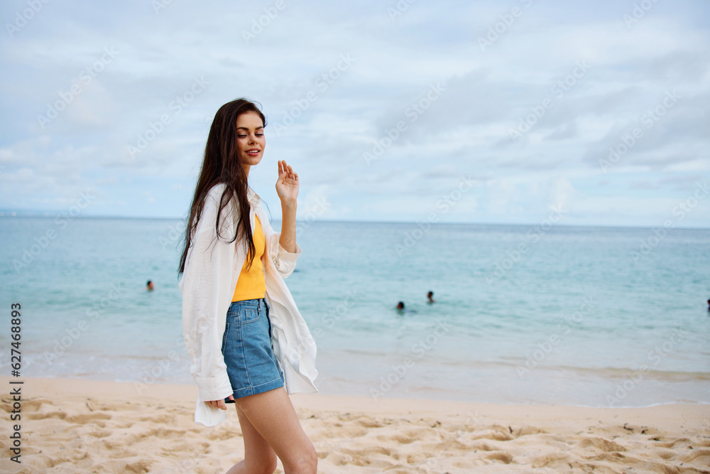 Sports woman runs along the beach in summer clothes on the sand in a yellow tank top and denim shorts white shirt flying hair ocean view, beach vacation and travel freedom Sports woman runs along the beach in summer clothes on the sand in a yellow tank top and denim shorts white shirt flying hair ocean view, beach vacation and travel freedom