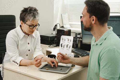 Man trying modern compact hearing aids at clinic. Audiologist consulting at hearing rehabilitation clinic to select digital device for middle age man. Doctor consulting patient photo