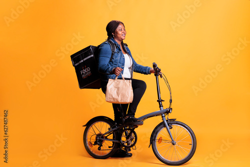 African american delivery woman holding paper bag while delivering lunch order to client using bike as transportation. Restaurant courier carrying takeout thermal backpack, takeaway service concept