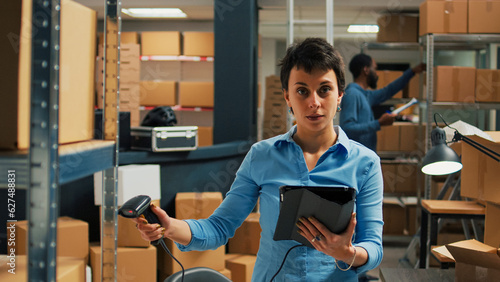 Young entrepreneur using scanner and tablet to do inventory, scanning bar code from packages on warehouse shelves. Woman checking stock of merchandise in boxes, supply chain logistics.