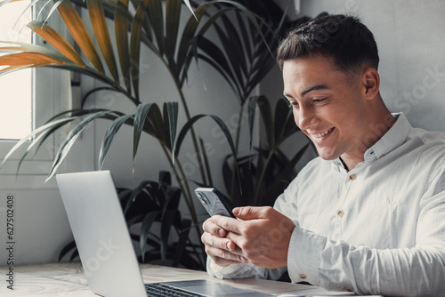 Confident man sitting at desk taking break in work with electronic documents on laptop to make answer telephone call. Smiling young guy freelancer synchronize data between home computer and smartphone