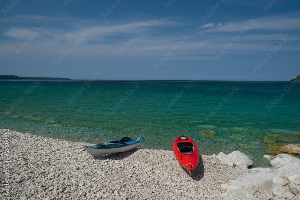 Two bright colorful kayaks on the white rocky beach by crystal clear see thru turquoise water. Sunny day, blue sky. Bruce Peninsula, Georgian Bay, Ontario, Canada.